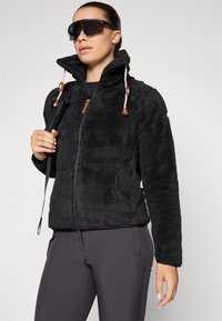 Femme portant une veste polaire noire à col haut, des leggings foncés, de grandes lunettes de soleil teintées, tenant la sangle d'un sac à dos sur son épaule.