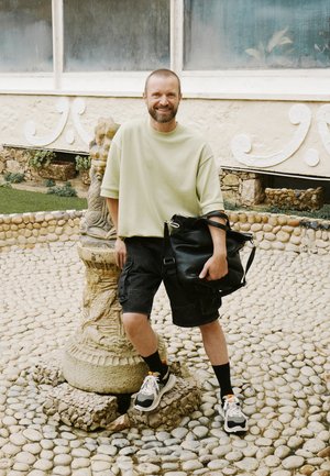 Borsa in pelle nera, camicia over-size verde chiaro, pantaloni cargo scuri, sneakers bianche con dettagli arancioni, in piedi accanto a un pilastro di pietra su un terreno di ghiaia.