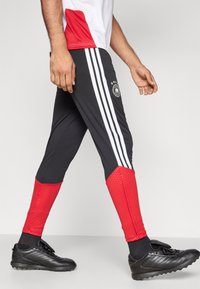 Sorte sportbukser med røde accenter og hvide striber, med et logo og teksturerede områder; båret sammen med sorte atletik sko.