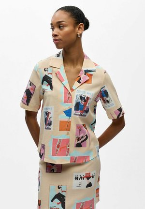 Frau trägt beige kurzärmeliges Hemd mit Knöpfen und Rock mit bunten abstrakten Postkarten- und Figurenmotiven, schaut seitlich und hat die Hände hinter dem Rücken.