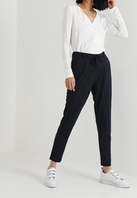 Donna che indossa una maglietta bianca a maniche lunghe, pantaloni neri con coulisse e sneaker bianche con velcro, in piedi contro uno sfondo chiaro e semplice.