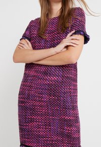 Šaty s krátkým rukávem vyrobené z texturované tkaniny, která má fialový a růžový tkaný vzor s třásněmi na rukávech a lemu.