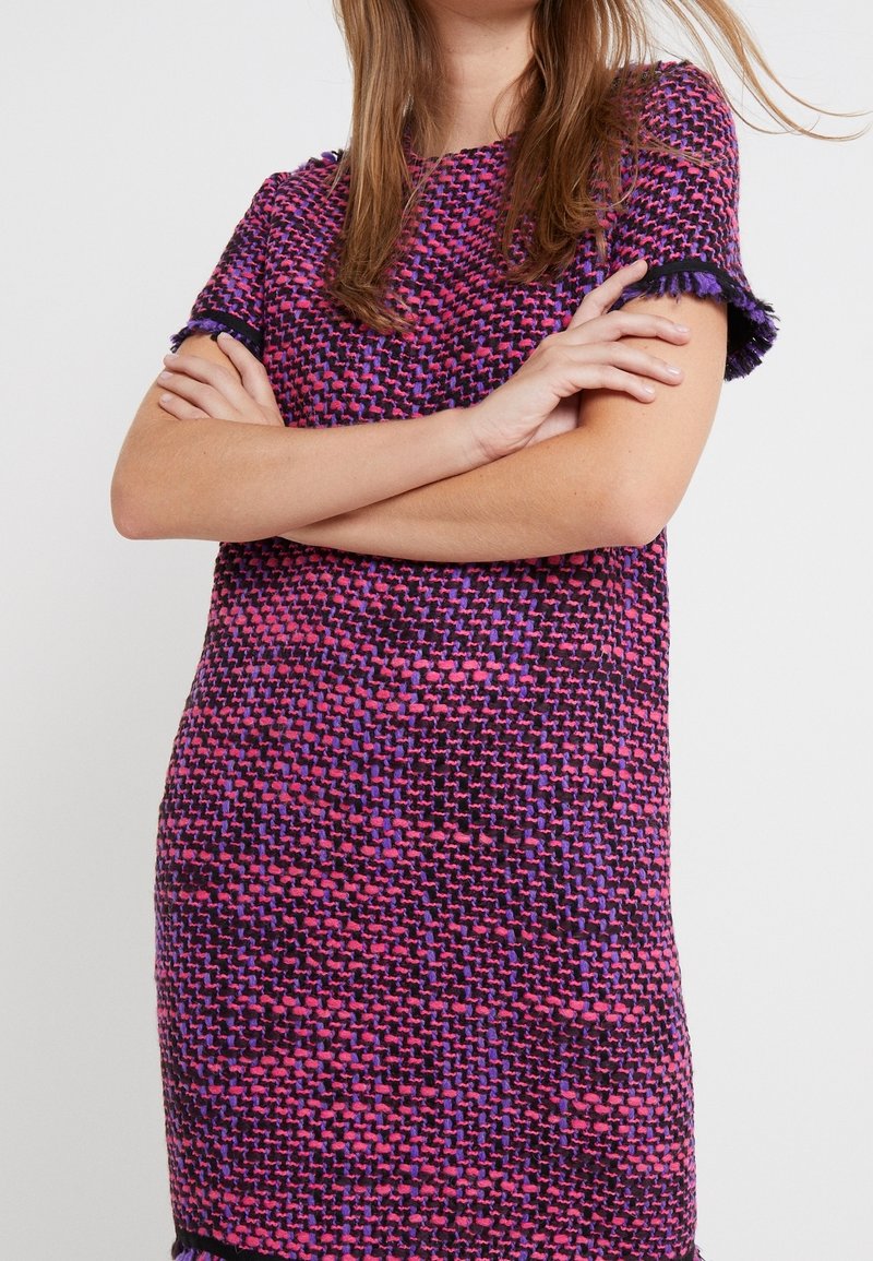 Šaty s krátkým rukávem vyrobené z texturované tkaniny, která má fialový a růžový tkaný vzor s třásněmi na rukávech a lemu.