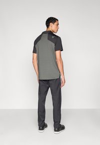 Κοντομάνικο πόλο σε ελιά με μαύρα πάνελ στους ώμους, συνδυασμένο με σκούρο γκρι παντελόνι και μαύρα παπούτσια, με διακριτική λεπτομέρεια λογότυπου.