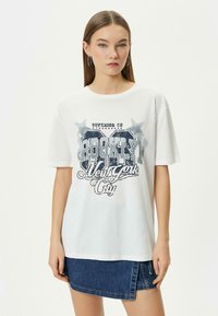 Camiseta blanca de algodón con un estampado gráfico azul que incluye estrellas y el texto: "BROOKLYN New York City." Corte holgado y mangas cortas.