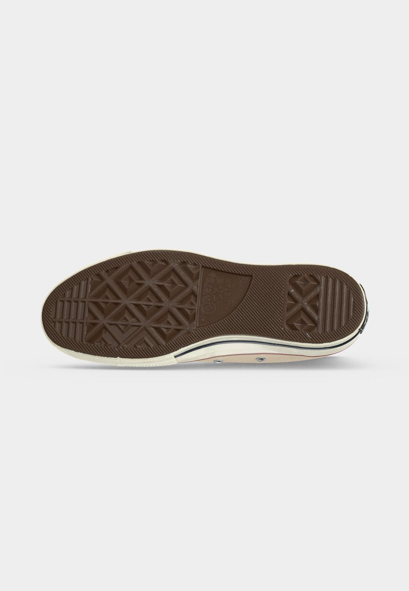 Braune Gummisohle eines Sneakers mit Rauten- und Linienprofil, sichtbarem weißen Rand und Teil des Oberschuhs mit Metallösen.