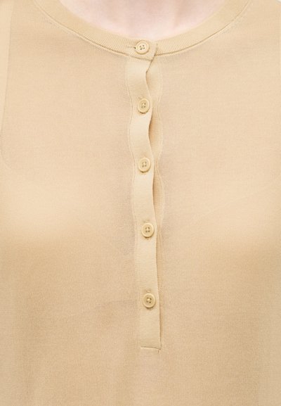 Κοντινή λήψη μπεζ πλεκτού τοπ με στρογγυλή λαιμόκοψη και έξι κατακόρυφα περασμένα κουμπιά στο κέντρο μπροστά.
