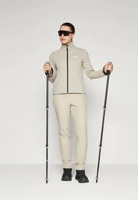 En person iført beige treningstøy og svarte turstøvler holder svarte turstaver, og står mot en enkel lys bakgrunn.