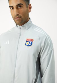 Hellgraue Sportjacke mit hohem Kragen, Reißverschluss, drei weißen Streifen auf der Schulter und einem gestickten Olympique Lyonnais-Logo.