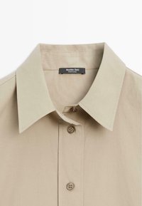Béžová košile na knoflíky s klasickým límečkem, dvěma předními knoflíky a látkovou nášivkou s nápisem "Massimo Dutti." Hladká textura, přizpůsobený design.