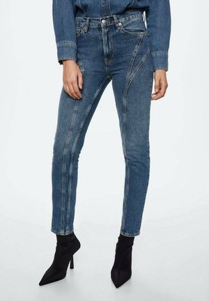 Kobieta nosząca niebieskie jeansy z wysokim stanem z unikalnymi ukośnymi szwami, w połączeniu z czarnymi botkami na obcasie z zaostrzonym noskiem.