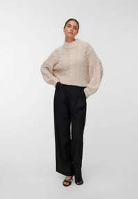 Femme portant un pull en laine beige à mailles, un pantalon large noir, des talons hauts noirs à lanières, se tenant avec les mains dans les poches devant un fond blanc.