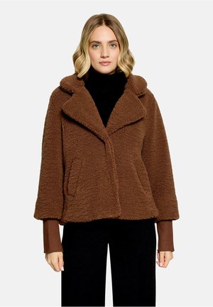 Woman with blonde hair wearing a brown fuzzy coat over a black top, standing against a plain light background.