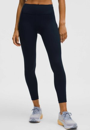 Woman wearing black high-waist leggings and light grey athletic shoes standing against a plain background.