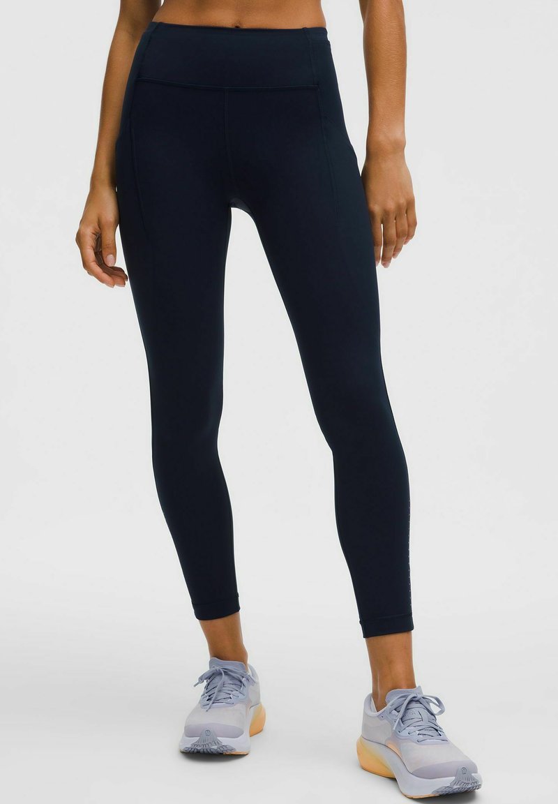 Egy nő fekete magas derekú leggingset és világosszürke sportcipőt viselve áll egy sima háttér előtt.