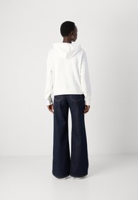 Kvinna som står vänd bortåt och har på sig en vit hoodie, mörka vida jeans och hoop-örhängen mot en enkel ljus bakgrund.