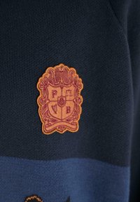 Wełniany sweter w kolorze granatowym z wyraźną wytłoczoną naszywką herbu w złotym i burgundowym kolorze, z inicjałami P, B oraz szczegółowym wzorem na bordzie.