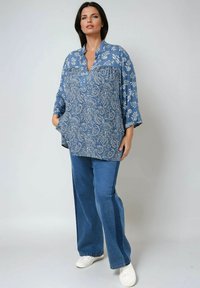 Woman standing wearing a blue paisley blouse with floral sleeves, wide-leg blue jeans, and white sneakers, against a plain background.