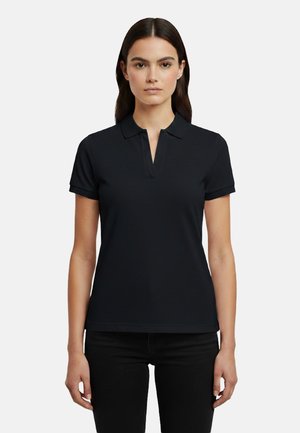 Giovane donna con capelli castani lisci, che indossa una polo nera a maniche corte e pantaloni neri, in piedi contro uno sfondo chiaro e semplice.