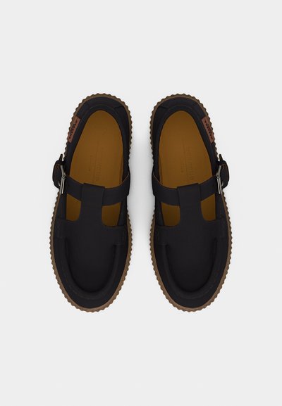 Ζευγάρι μαύρα παπούτσια στιλ Mary Jane με καφέ σόλες και ασημένιες αγκράφες, φωτογραφημένα από πάνω σε λευκό φόντο.