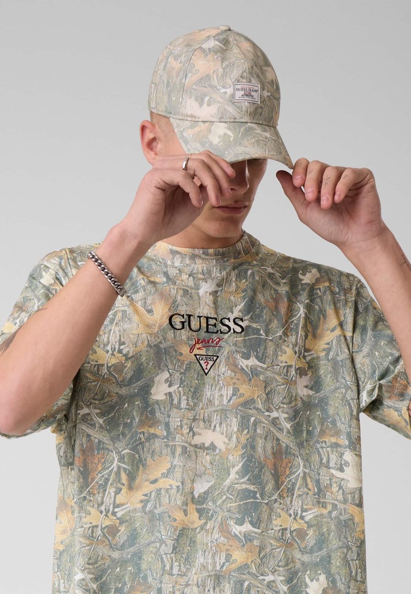 Jeune homme ajustant une casquette à motif camouflage et portant un t-shirt camouflage assorti sur un fond gris uni.