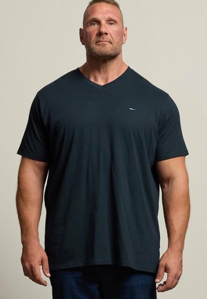 Muskelbepackter Mann mit kurzen Haaren und Bart, der ein schlichtes schwarzes T-Shirt mit V-Ausschnitt trägt, vor einem beigen Hintergrund stehend.