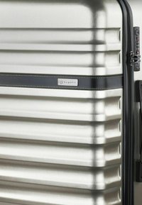 Valigia rigida argentata con scanalature verticali, striscia di accento nera, etichetta con marchio e serratura a combinazione sul lato. Finitura metallica liscia.