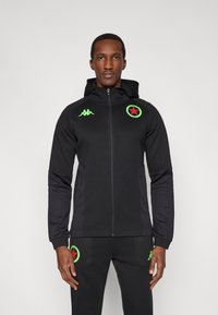 Čierna mikina na zips s kapucňou, zeleným logom hviezdy na hrudi a zodpovedajúcimi zelenými detailmi. Mäkká látka, priliehavý strih s bočnými vreckami.