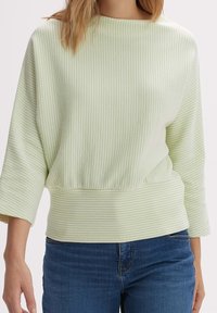 Femme portant un pull côtelé à manches longues vert clair avec de larges poignets et un jean en denim bleu, debout devant un fond uni.