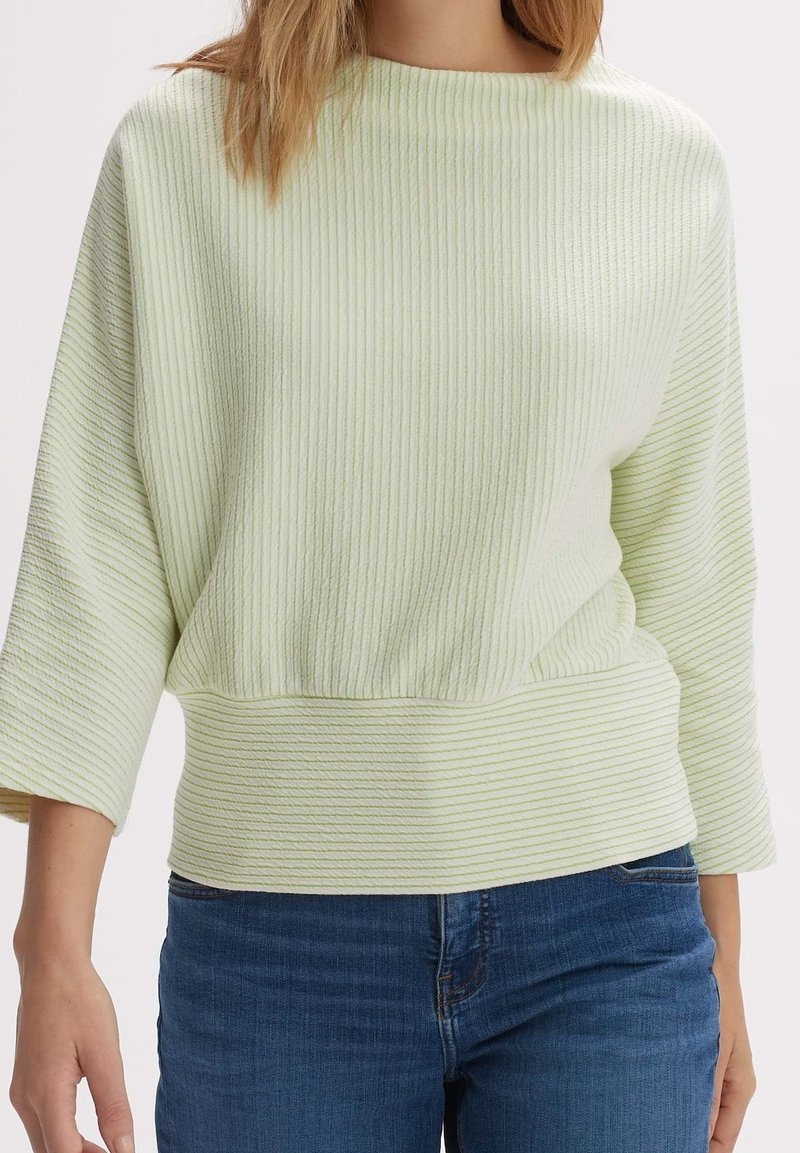 Femme portant un pull côtelé à manches longues vert clair avec de larges poignets et un jean en denim bleu, debout devant un fond uni.