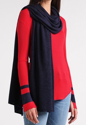 Femme portant un pull rouge côtelé avec des rayures bleu marine sur les manches et une longue écharpe bleu marine drapée sur les épaules, associée à un jean bleu clair.