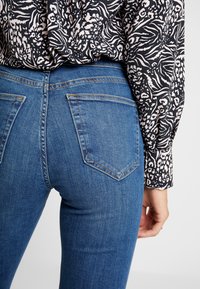 Siniset farkut, joissa on istuva malli ja takataskut sekä kontrastivärinen ompelu, yhdistettynä pitkähihhaiseen musta-valkoiseen kuvioituun paitaan.