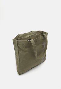 Sac fourre-tout en toile vert olive avec deux longues poignées, de forme rectangulaire et impression discrète du logo sur le côté. Dispose d'une ouverture sur le dessus pour un accès facile.