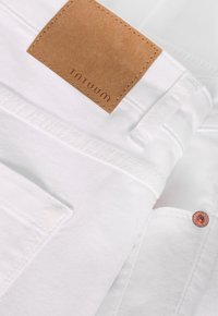 Vita denimjeans med en beige läderlapp märkt "TNTUUM", med kopparfärgad hårdvara och ren sömnad.