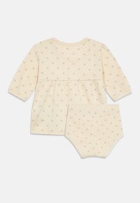 Conjunto de roupa em algodão em bege claro, com mangas longas, cintura franzida e parte inferior correspondente. Apresenta uma pequena impressão texturizada por todo o conjunto.