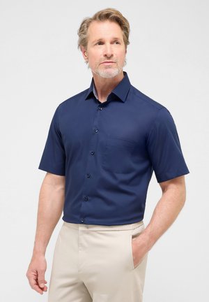 Marineblaues Kurzarm-Hemd mit Knopfleiste, einer Brusttasche und einem spitzen Kragen, das eine subtile Textur und schwarze Knöpfe aufweist. Kombiniert mit beigen Hosen.