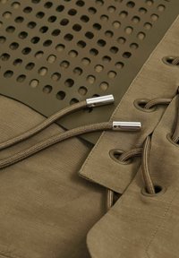 Olijfgroen stof met grote perforaties, met gestructureerd materiaal en zilverkleurige hardwaredetails, waaronder koord en oogjes.