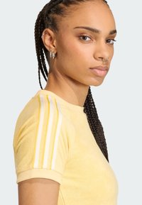 Žltá košeľa s krátkym rukávom vyrobená z textúrovaného materiálu, s bielymi a žltými pruhmi na pleciach. Detailný záber profilového pohľadu modela.