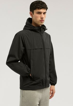 Junger Mann trägt eine schwarze Kapuzenjacke mit Reißverschluss und elastischen Bündchen sowie hellgraue Hose, steht vor einem einfarbigen Hintergrund.