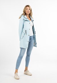 Lichtblauwe waterdichte jas met een capuchon, drukknoopsluitingen, zijzakken en trekkoordaccenten, gecombineerd met lichtgekleurde jeans en beige sneakers.