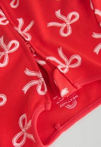 Indumento rosso con zip, caratterizzato da un motivo di fiocchi bianchi. Tessuto morbido con bordi a costine e un'etichetta con le istruzioni per la cura.