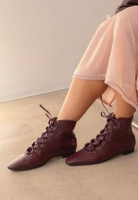 Botas de tobillo de cuero burdeos con punteras afiladas, que presentan un diseño de cordones decorativos y suelas planas, usadas con una falda rosa claro.