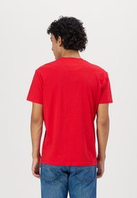 T-shirt en coton rouge avec col rond, manches courtes et un design de dos simple. Porté avec un jean en denim bleu montrant les poches arrière.