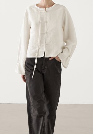 Person wearing a loose white long-sleeve top with front ties and black high-waisted relaxed pants, hand in pocket.