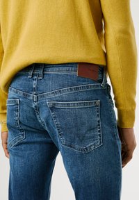 Jean en denim bleu avec une finition légèrement délavée, présentant des poches arrière et une étiquette de marque en cuir brun. Porté avec un pull jaune.