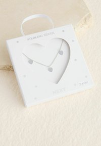 Náhrdelník zo striebra sterling s tromi malými príveskami v tvare srdca; zabalený v bielom krabičke s okienkom v tvare srdca a dizajnom s bodkami.