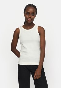 Weißes ärmelloses Tanktop mit geripptem Texture und kontrastierendem Ausschnitt. Das Model steht selbstbewusst da, mit entspannten Armen an ihrem Körper.