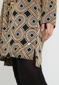 Beige und schwarze gemusterte Stoff mit geometrischen Designs. Seitenschlitz-Detail und kleine Metallakzent am Saum. Glatte Textur.