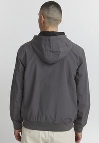 Giacca leggera grigia con cappuccio, orlo a costine, texture liscia e polsini elastici. Presenta un design semplice e funzionale adatto per essere indossato a strati.