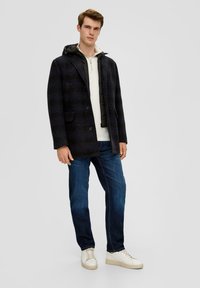 Marineblauwe geruite jas met knoopsluiting en capuchon, gelaagd over een lichtgekleurde trui, gecombineerd met donkere jeans en witte sneakers.